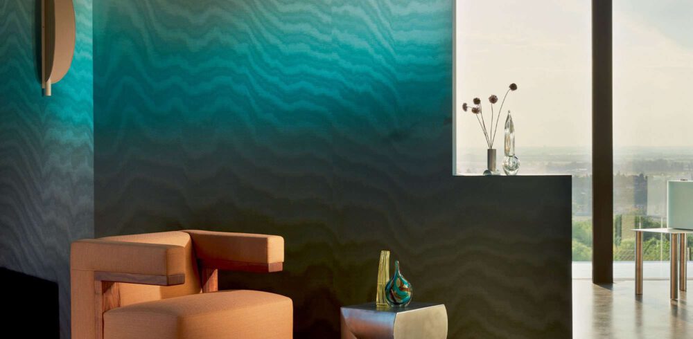 Großzügige Moire Tapete auf Vlies, türkis-blauer Farbverlauf, davor ein cognac-farbener Cocktail chair